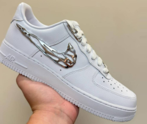 White Nike Air Force 1 sneaker with a shiny metal chain swoosh held in a hand against a neutral background