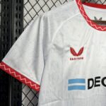 White sports jersey with red wing Castore logo on chest and red zigzag trim at sleeve edge, hanging on a black metal rack behind a chain-link fence.