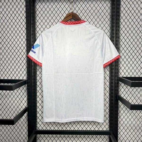 White soccer jersey hanging on a wooden hanger, red zigzag trim at the collar and sleeve cuffs against a metal mesh backdrop.