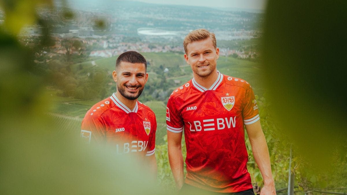 Two male soccer players in red jerseys smile for the camera against a hillside town backdrop with blurred greenery in the foreground.