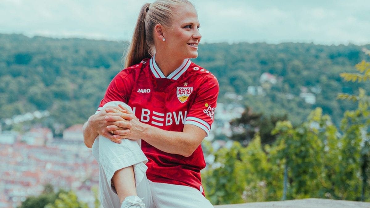 Woman in a red sports jersey holding a young child in white pants outdoors, with a blurred hillside town in the background.