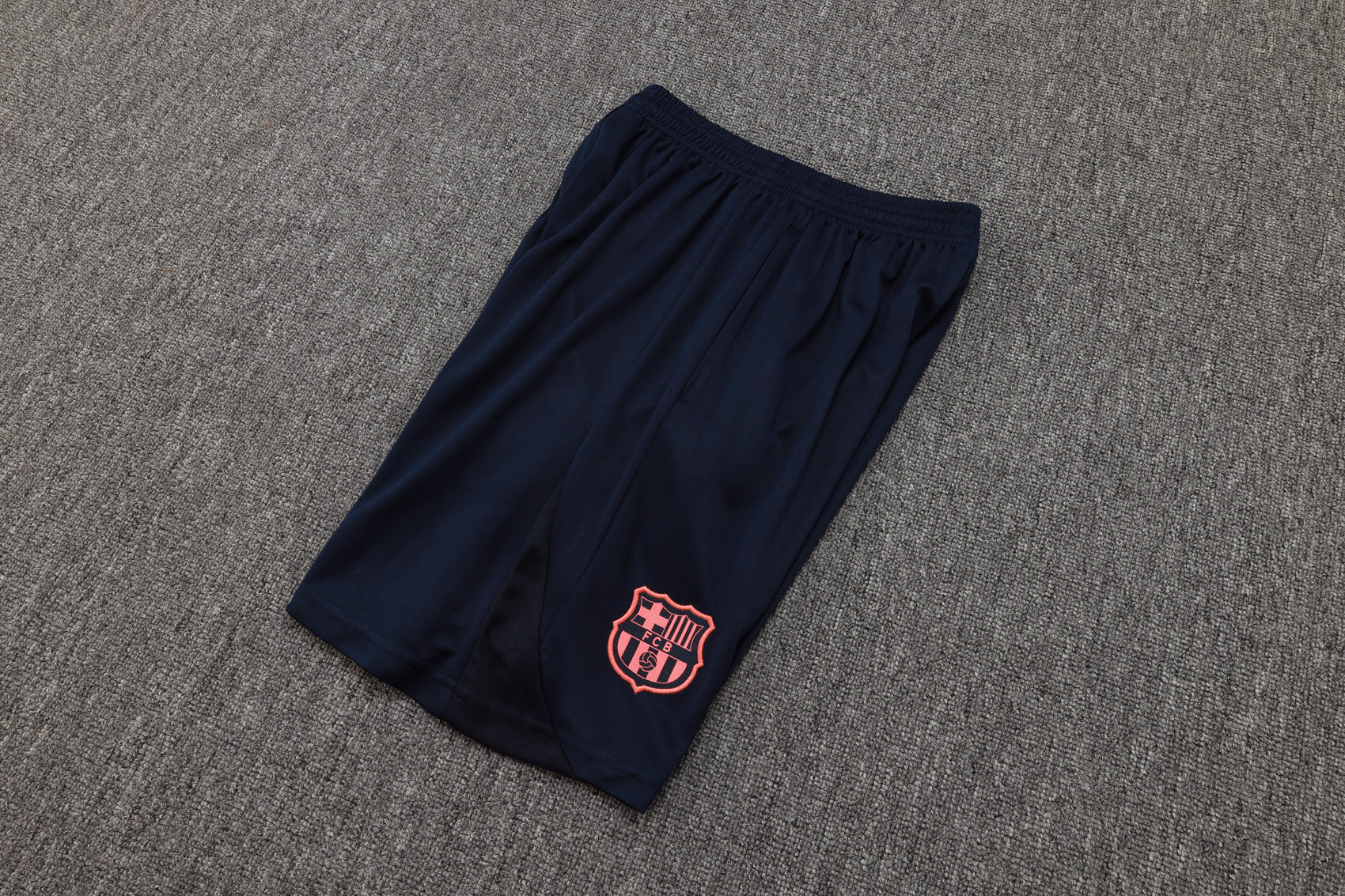 202c12c2 | UAE SPORT STORE Navy athletic shorts with a pink FC Barcelona crest on the lower leg, laid on a gray carpeted floor.