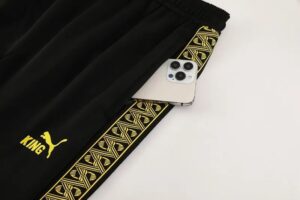 Black athletic pants with a yellow patterned stripe and Puma 'KING' logo, a white iPhone tucked into the side pocket.