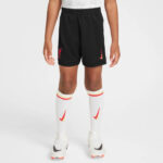 Child wearing black athletic shorts with a red club crest on the left, white socks with red/yellow stripes, and white soccer cleats with a black swoosh.