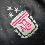 Close-up of a white shield patch with pink ALA letters, pink stripes, and a laurel border on a dark speckled fabric background.