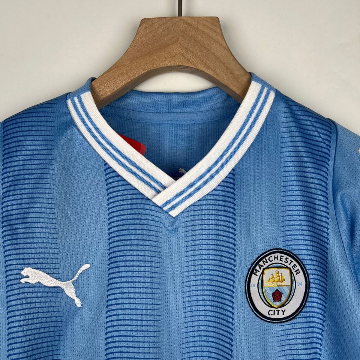 Blue Manchester City football jersey hanging on a wooden hanger against a plain wall, close-up of collar and crest visible