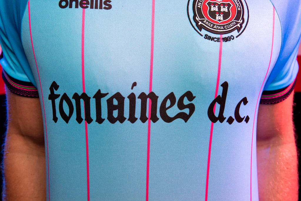 2124CFC8-BD19-4CEA-A808-C78AAAAB90EE | UAE SPORT STORE Light blue rugby jersey with pink vertical stripes and the text 'fontaines d.c.' across the chest, plus a crest on the upper right side of the shirt.