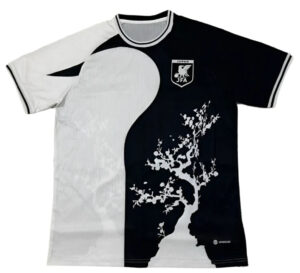 Split-color soccer jersey: white left, black right, featuring a white tree graphic on the dark side and a JFA crest on the chest area (Japan).