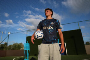 Male soccer player in Chelsea training kit holds a ball on a practice pitch with a goal and net in the background.