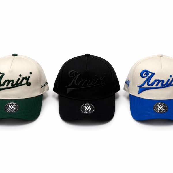 Three Amiri baseball caps: cream with green brim, black, and cream with blue brim; 'Amiri' script on the front edge and a circular patch on the bill area.