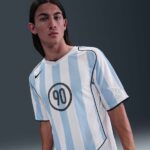 Young man wearing a light blue and white striped soccer jersey with the number 90 on chest, posing against a dark background.