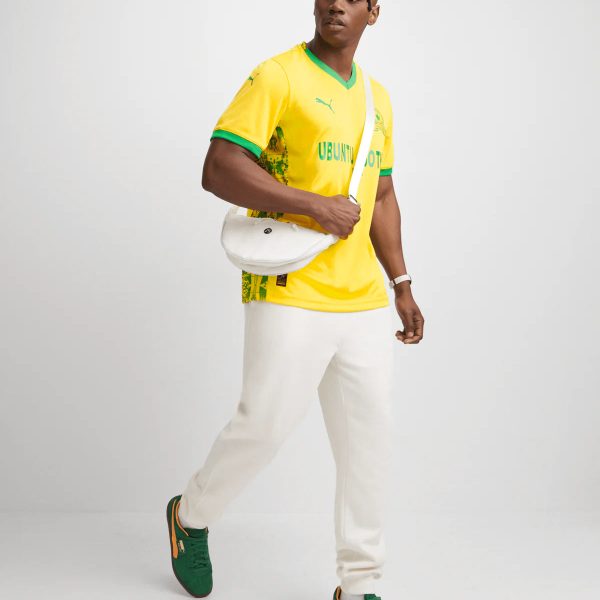 Man wearing a bright yellow jersey with green trim, white pants, and green sneakers, carrying a white crossbody bag against a white background.