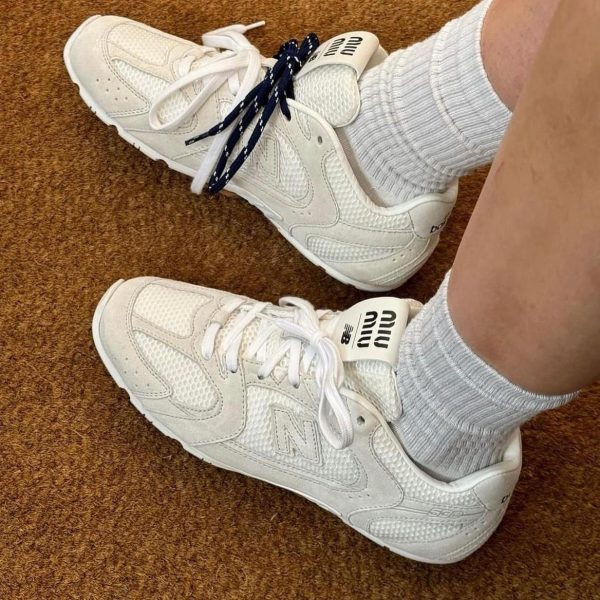 White New Balance sneakers on a brown carpet, with gray socks visible, one shoe laced in navy more than the other.