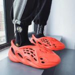 Bright orange slip-on sandals with cutouts worn over black socks by a window.