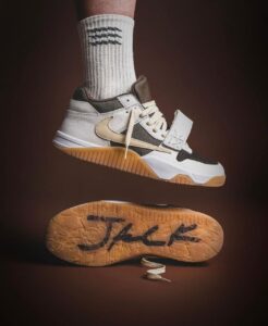 Nike sneaker in white, beige, and black with a gum sole, suspended mid-air over a textured outsole on a brown background.