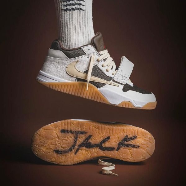 Nike sneaker in white, beige, and black with a gum sole, suspended mid-air over a textured outsole on a brown background.