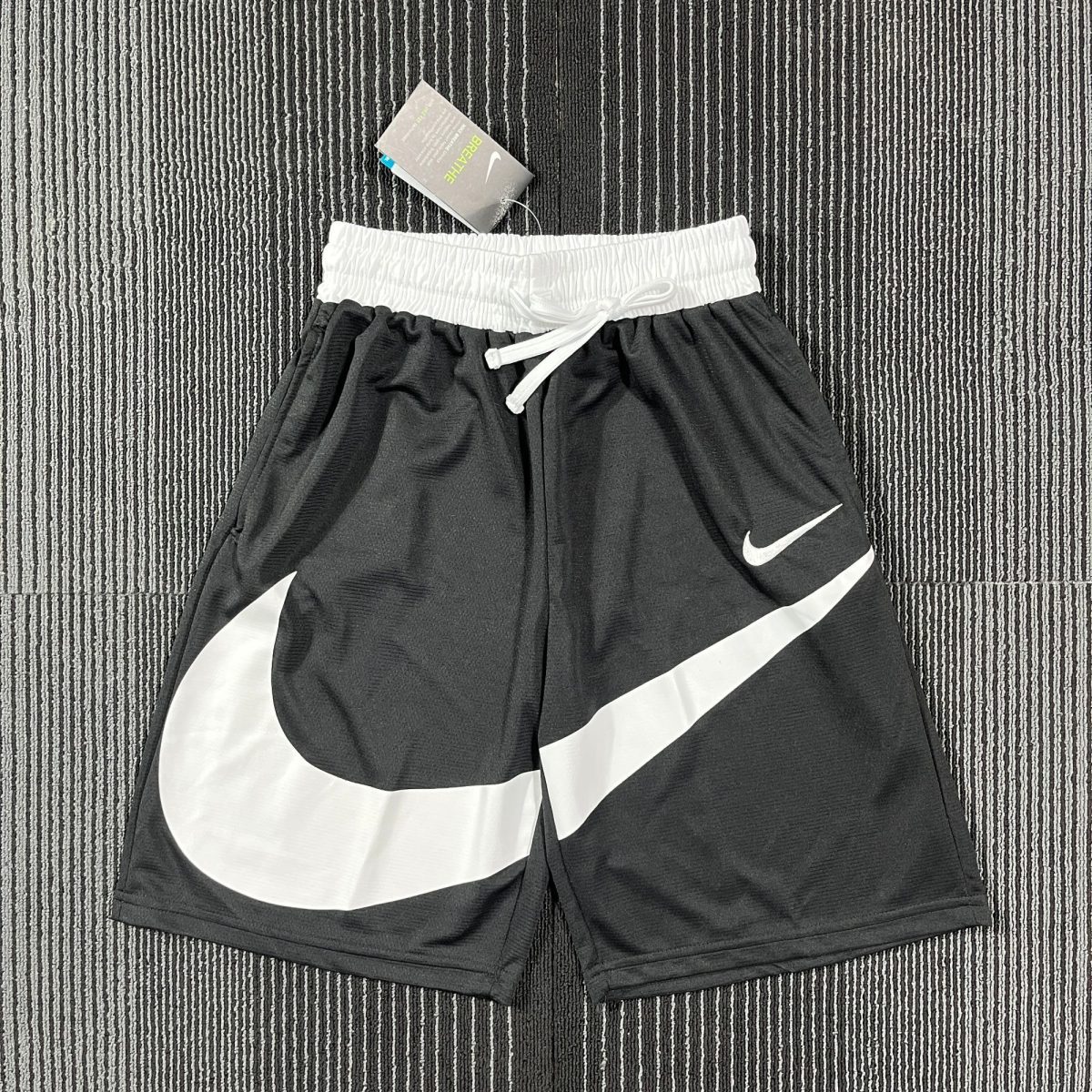 Black athletic shorts with a white elastic waistband and Nike swoosh logos on the legs, laid on a striped carpet.