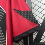 Close-up of red fabric with a black diagonal band and white stitched edge, with a chain-link fence in the background.