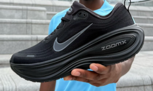 Black Nike ZoomX running shoe held by a person in a blue shirt, showing the Nike swoosh against a stair background.