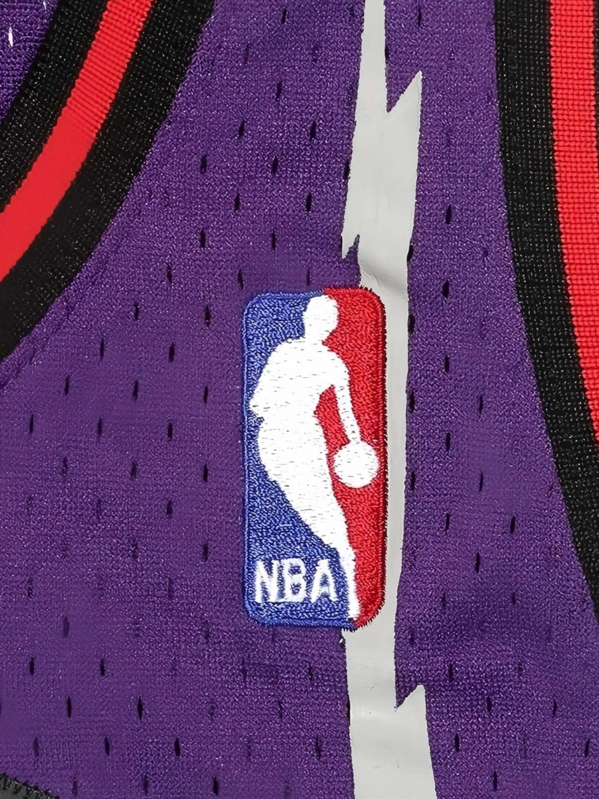 Close-up of the NBA logo embroidered on purple athletic fabric with a white seam and red/black stripes nearby.
