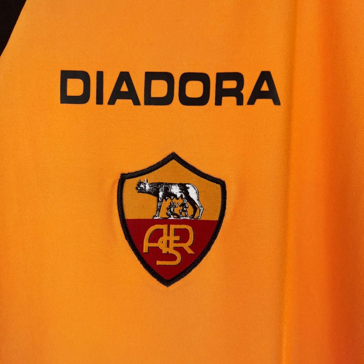 Close-up of orange fabric with bold black letters spelling 'DIABORA' over a shield emblem partially visible underneath.