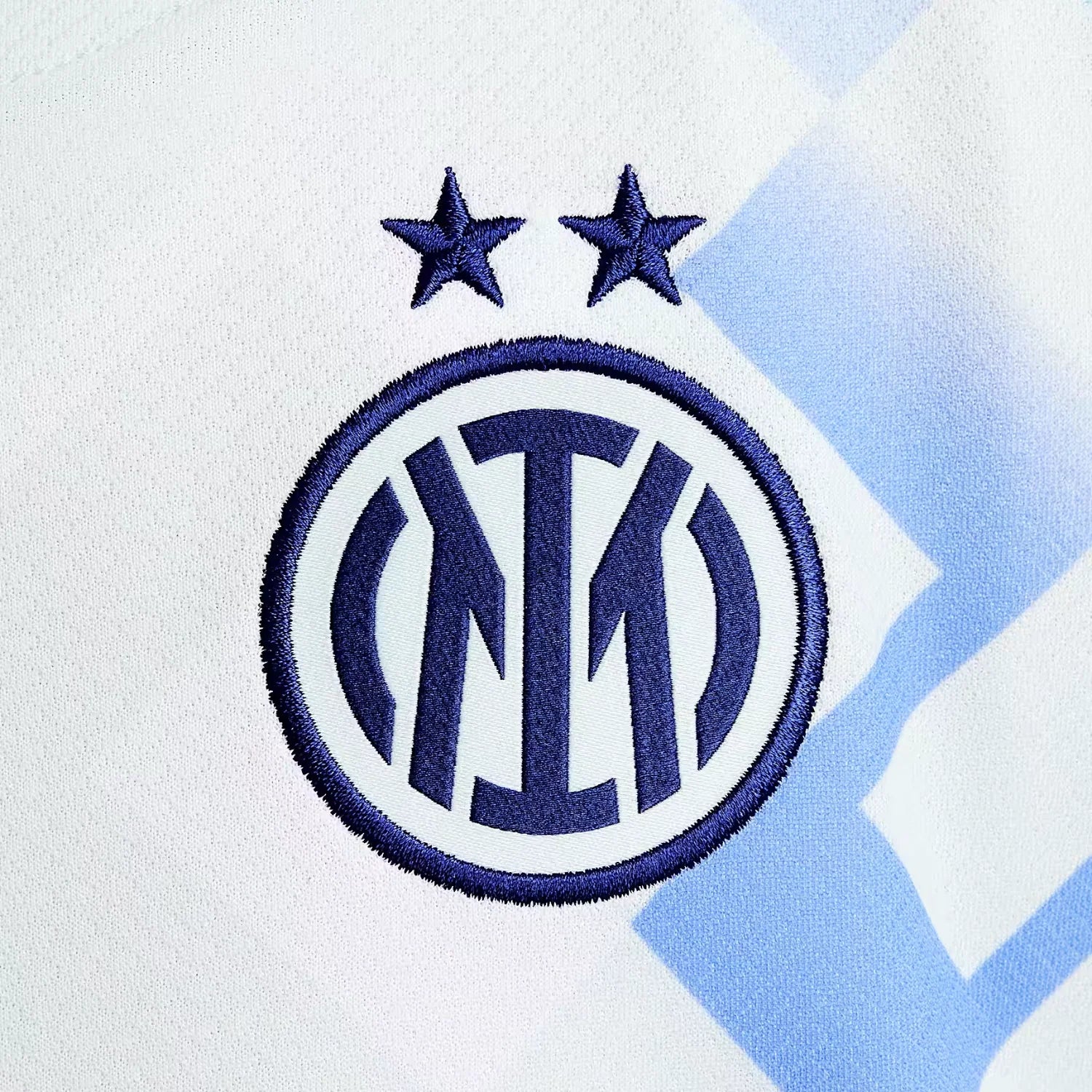 22_b4f87345-99c9-46b0-ac17-d7910c022e2c | UAE SPORT STORE Close-up of white fabric featuring a blue embroidered Inter Milan crest with two stars above the circle.