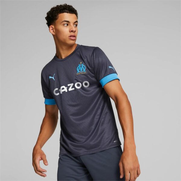 Soccer player in a dark blue training shirt with light blue sleeve cuffs, looking to the left against a gray background.