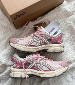 Pair of pink and white ASICS Gel-Kayano sneakers with matching laces on a white patterned surface, box in the background.