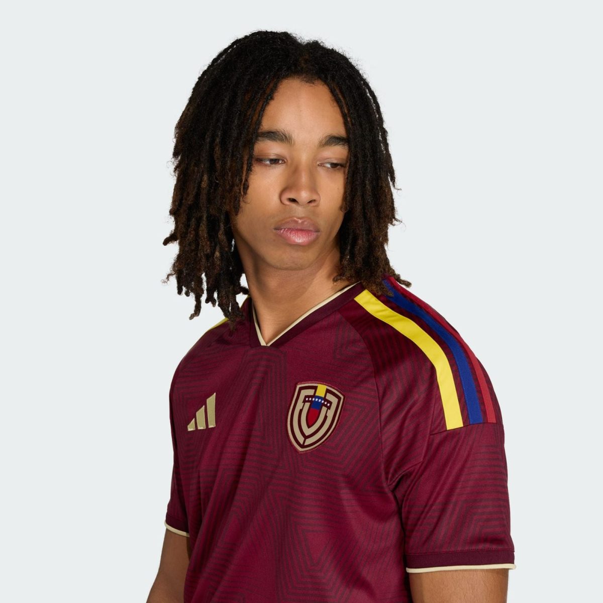 Young man with dreadlocks wearing a maroon soccer jersey with yellow and blue shoulder stripes, looking to the side.
