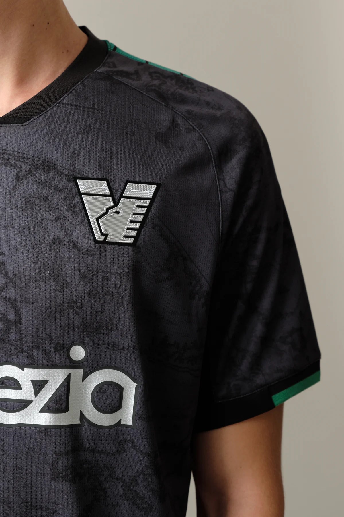 23_Home_Jersey_Venezia_4_5c199846-f548-47bf-843e-0c605f06e0fc | UAE SPORT STORE لقطة مقرّبة لقميص رياضي أسود مع شعار EA7 فضي على الصدر وحافة خضراء على الكم.