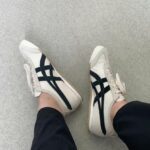 23e27621-41cc-4cf3-be09-c3c07f34b212 | UAE SPORT STORE White sneakers with black side stripes worn by a person seated with legs crossed on a light floor surface.