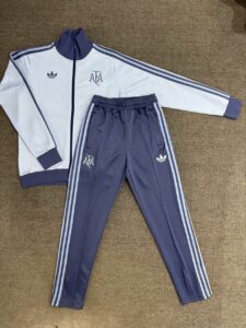 Adult-sized sports tracksuit laid flat: white zip-up jacket with navy cuffs and three navy stripes along sleeves, matching navy pants with white side stripes and Adidas logos. AFA crest on jacket and leg.