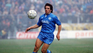 Soccer player in a blue uniform controls a ball on a grassy field during a match, with a blurred crowd in the background.