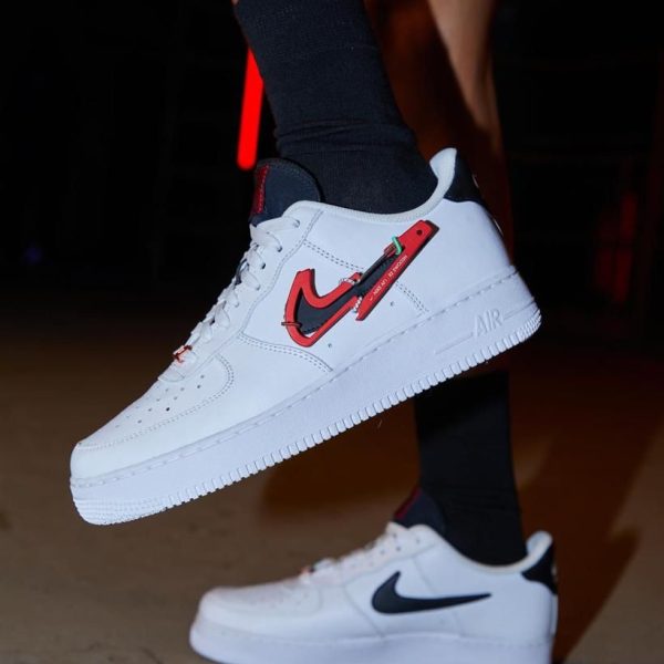 White low-profile Nike sneakers with a black swoosh and red accent tag, being shown mid-air by a wearer in black socks.