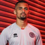 Portrait of a man with a short beard wearing a white Sheffield United FC jersey, posed against a red striped background.