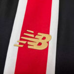 Close-up of a gold New Balance 'NB' logo on a red stripe with white edges against a dark background, cropped diagonal.
