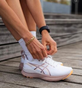 Close-up of a person tying white sneakers with peach soles on a wooden deck, wearing white socks, a gold bracelet, and a black smartwatch.