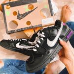 Black Nike sneakers with white swoosh held in hand; a shoebox with circular cutouts nearby, and a person wearing ripped jeans.