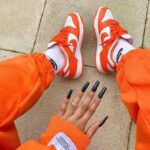 Top-down view of orange-and-white sneakers, white socks, and orange joggers with a hand showing long black nails nearby.