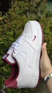 Close-up of a white Nike Air Force 1 sneaker with burgundy accents being held by a hand, with greenery in the background.