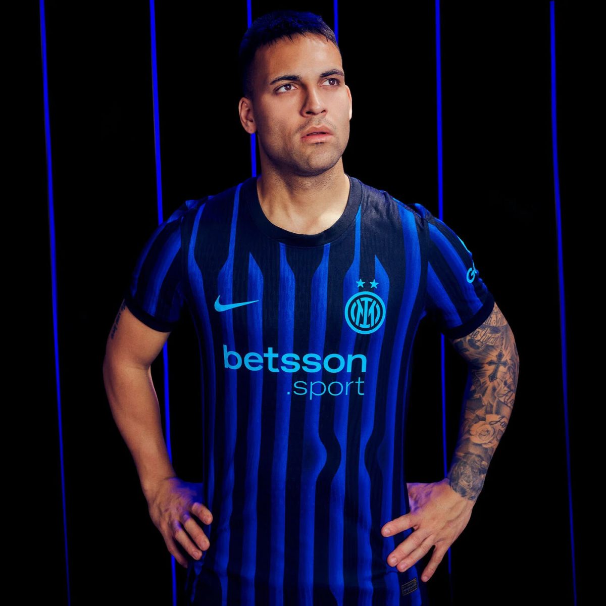 Man standing with hands on hips wearing a blue and black Inter Milan jersey featuring the Betsson sponsor and crest, against a dark background with blue vertical lines.