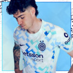 Male model wearing an Inter Milan white and blue geometric soccer jersey, looking down against a blue background.