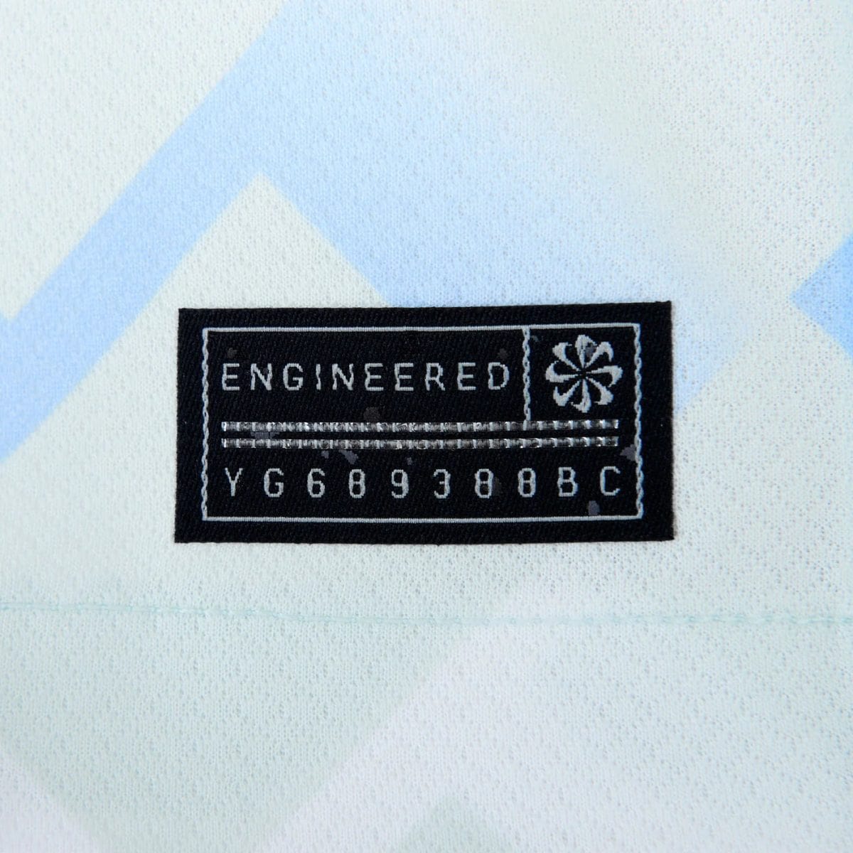 Black sewn label reading 'ENGINEERED' with a small emblem on the right and a row of numbers underneath, sewn onto light fabric