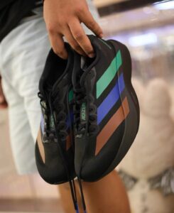 Black sneaker with multicolored diagonal stripes (green, blue, orange) held by a hand.