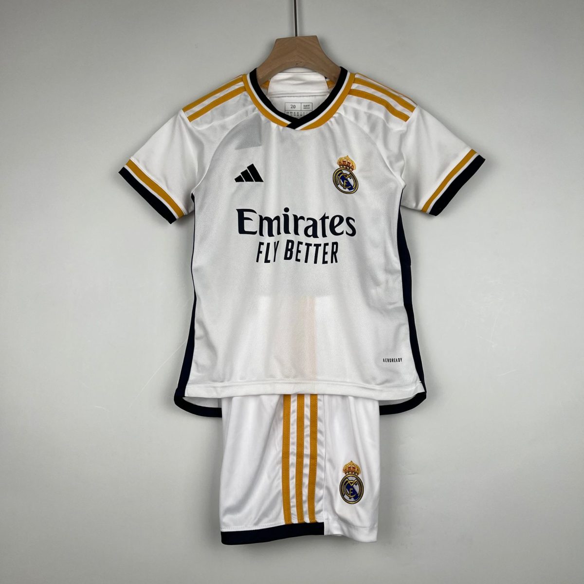 White Real Madrid jersey on a wooden hanger with gold stripes, featuring Adidas logo, club crest, and 'Emirates Fly Better' sponsor text.