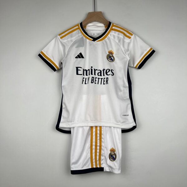 White Real Madrid jersey on a wooden hanger with gold stripes, featuring Adidas logo, club crest, and 'Emirates Fly Better' sponsor text.