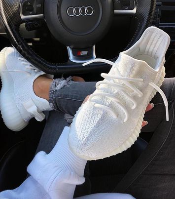 White knit sneakers resting on a person's lap inside a car, with an Audi steering wheel visible in the background.