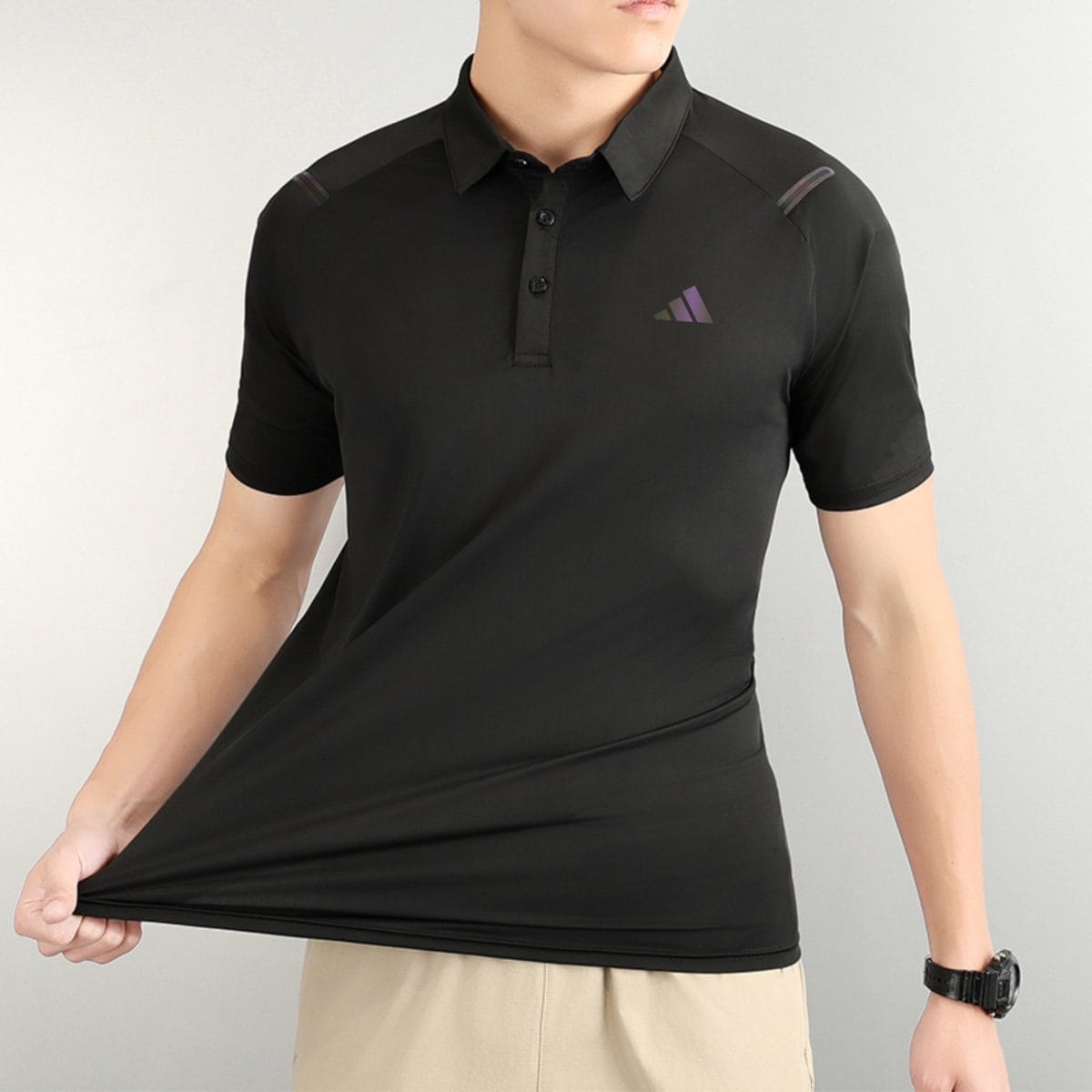 Person wearing a black polo shirt with a small purple triangle logo on the chest, beige shorts, and a black wristwatch visible on the right wrist.