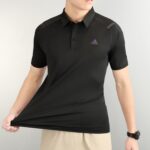 Person wearing a black polo shirt with a small purple triangle logo on the chest, beige shorts, and a black wristwatch visible on the right wrist.