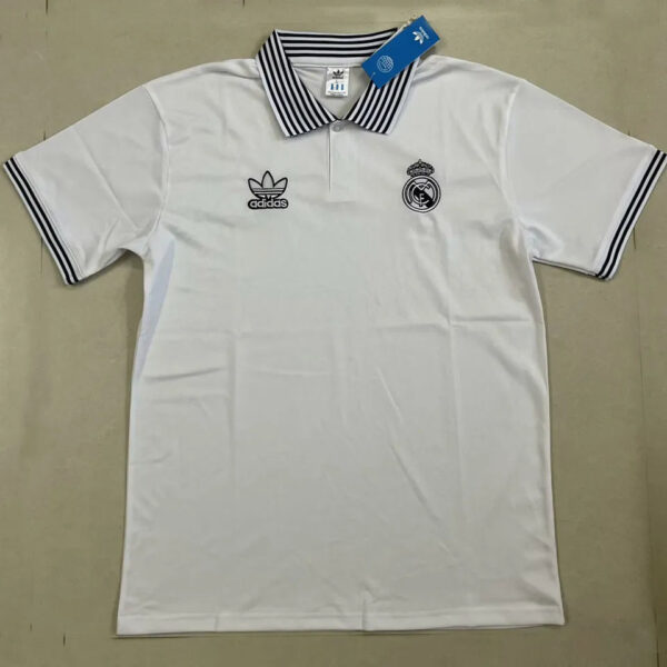 White polo shirt with black-striped collar and sleeve cuffs, Real Madrid crest on the left chest and Adidas logo on the right chest with a blue tag attached (brand product).